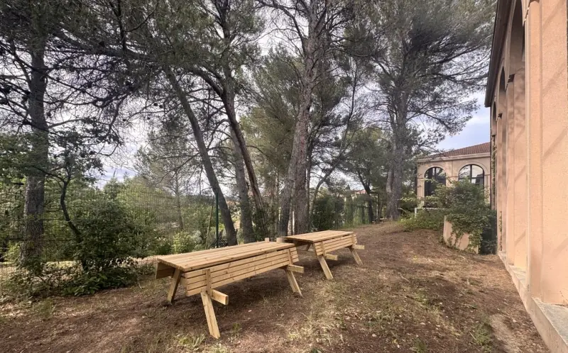 Tables de pique-nique en bois sous les pins, au pied du bâtiment à arcades